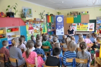 Na pierwszym planie siedzi grupa dzieci, na drugim planie widać kobietę opowiadającą bajkę, w tle wnętrze sali przedszkolnej oraz baner Punktu Informacji Europejskiej Europe Direct w Olsztynie.