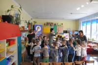 Na pierwszym planie grupa dzieci i nauczycielek, bawiąca się w pociąg, w tle wnętrze sali przedszkolnej oraz baner Punktu Informacji Europejskiej Europe Direct w Olsztynie.