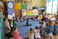 Na pierwszym planie grupa dzieci siedząca w kręgu i słuchająca prowadzącej, która trzyma w rękach mapę Wielkiej Brytanii i zadaje pytanie, jedno z dzieci podnosi rękę w odpowiedzi. W tle wnętrze sali przedszkolnej oraz baner Punktu Informacji Europejskiej Europe Direct w Olsztynie.