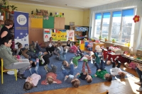 Na pierwszym planie grupa dzieci leżąca na podłodze z podniesionymi nogami podczas zabawy ruchowej, w tle wnętrze sali przedszkolnej oraz baner Punktu Informacji Europejskiej Europe Direct w Olsztynie.