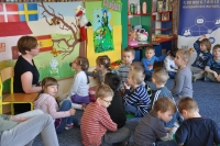 Na pierwszym planie grupa dzieci siedząca w grupie i słuchająca prowadzącej, która siedzi na tle mapy Hiszpanii zawieszonej na ścianie przedszkola. W tle wnętrze sali przedszkolnej oraz baner Punktu Informacji Europejskiej Europe Direct w Olsztynie.