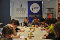 Na zdjęciu siedzący przy stołach i rysujący uczestnicy warsztatów, na stołach widoczne materiały plastyczne, w tle banery Wojewódzkiej Biblioteki Publicznej w Olsztynie i Punktu Informacji Europejskiej Europe Direct w Olsztynie.