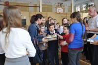 Na zdjęciu znajduje się po lewej stronie nauczycielka otoczona grupą dzieci. Nauczycielka trzyma w rękach pudełko z grą planszową. Po prawej stronie stoi jedna z osób prowadzących zajęcia.