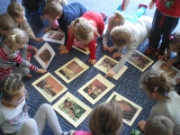 Grupka dzieci siedzi na podłodze tworząc mały okrąg. Między nimi, w środku koła na podłodze leża zdjęcia, które dzieci oglądają.