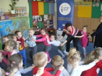 Zdjęcie przedstawia grupę przedszkolaków ustawionych w kółko. Dzieci bawią się w „pociąg”. W tle banner Europe Direct.
