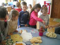 Na zdjęciu grupka przedszkolaków otacza stół na którym znajdują się owoce i fontanna. Dzieci nabijają owoce na wykałaczki i maczają w fontannie.
