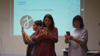 2019-10-02_Olsztyn_Aplikacje_8 - 3 uczestniczki  używają smartfonów (korzystają z aplikacji wskazanych przez trenera) - stoją na tle slajdu na ściennym ekranie z napisem : 
