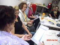 Zdjcie przedstawia 5 kobiet zgromadzonych przy stole z poczstunkiem. Kobiety piewaj piosenk spogldajc w tekst. W oddali po prawej stronie widzimy zbiry muzoteki, po lewej st z kosmetykami jednej z czytelniczek, ktra udzielaa porad.