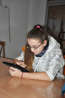 Na zdjęciu nastolatka, która siedzi przy stole. Trzyma  w rękach tablet oparty  o stół. Nastolatka patrzy  w tablet  i go obsługuje prawą ręką.