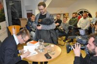 Na pierwszym planie Dawid Kraszewski. Poeta siedzi przy stole i podpisuje swoj ksik. W tle uczestnicy spotkania.