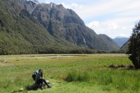 Zdjcie z wyprawy do Nowej Zelandii. Na pierwszym planie zielona polana. W lewym dolnym rogu na kamieniu siedzi Kornelia Tokarczyk. Na drugim planie masywna gra i bkitne niebo.