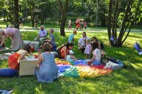 Strefa zabaw dla dzieci przygotowana przez Pitk - Bibliotek Dzieci i Modziey. Na wielokolorowej chucie siedz dzieci z rodzicami, dookoa park. 