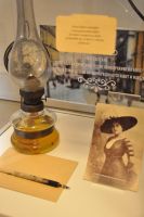 Fragment ekspozycji w szklanej gablocie: czarno-biae zdjcie Emilii Sukertowej w duym, zdobnym pirem kapeluszu oraz lampa naftowa.