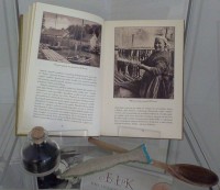 Fragment ekspozycji na jednej z pek: potwarta ksika z ilustracj przedstawiajc kobiet suszc ryby, drewniana yka, rybka uszyta z ptna oraz szklane naczynie z nasionami czarnuszki.