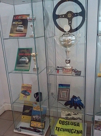 Dwie szklane witryny z widocznymi eksponatami na siedmiu plkach, m.in. z kierownic samochodu sportowego oraz srebrny puchar z zawodw oraz kolorowymi publikacjami na temat sportu samochodowego.