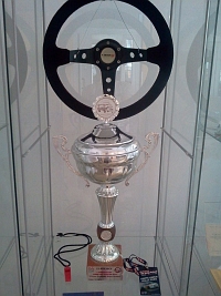 Fragment wystawy w witrynie: kierownica samochodu sportowego oraz duy, srebrny puchar z zawodw.