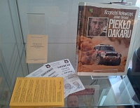 Fragment wystawy w witrynie: ustawione pionowo ksika „Pieko Dakaru” Krzysztofa Hoowczyca oraz Juliana Obrockiego i Statut Warmisko-Mazurskiego Klubu Automobilowego oraz pooone obok dwie licencje pilota samochodowego.