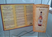 Fragment wystawy w witrynie: dwie ulotki lawendowego piwa rzekomej receptury Mikoaja Kopernika - jedna przyczepiona na bordowym tle kartonowej podstawki, druga ustawiona pionowo, z wizerunkiem butelki piwa na awersie.